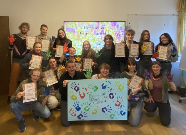 Eine Gruppe von fast 20 Personen zeigt sich mit ihren Zertifikaten und einem Poster in der Mitte mit ihren Handabdrücken in vielen Farben.