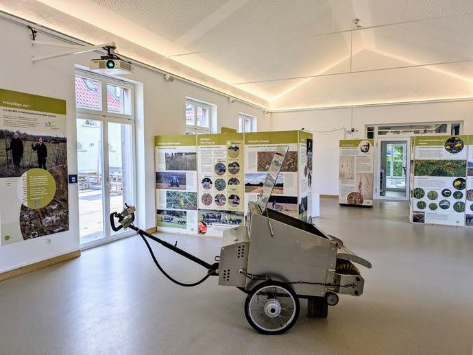Blick in den Ausstellungsraum "Wo die wilden Pflanzen leb(t)en" mit Tafeln und einem Wiesenmähgerät (vergrößerte Bildansicht wird geöffnet)